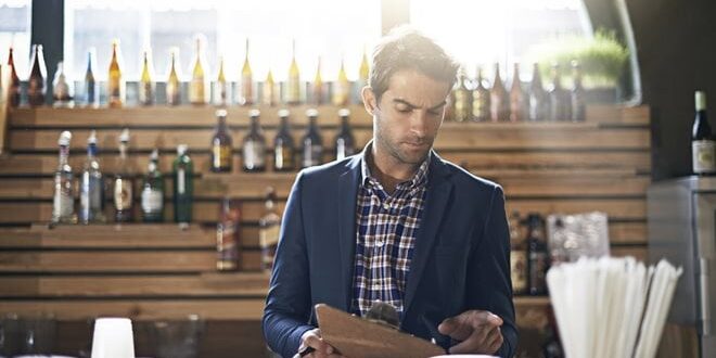 A man in a blazer looks at a clipboard behind a bar with bottles lining the shelves. White cups are in the foreground.