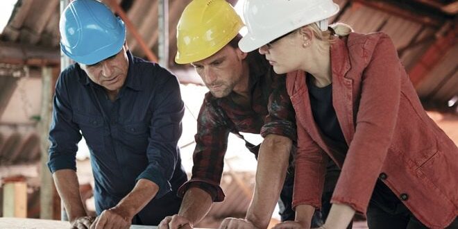Three construction workers in hard hats review plans on a table in an industrial setting.