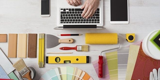 Hands typing on a laptop surrounded by design tools, color swatches, paintbrushes, a roller, a smartphone, and a tablet on a light wooden desk.