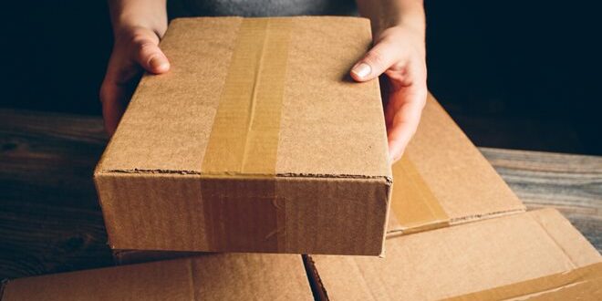 A person holding a brown cardboard box with packing tape on a wooden surface, surrounded by three similar boxes.