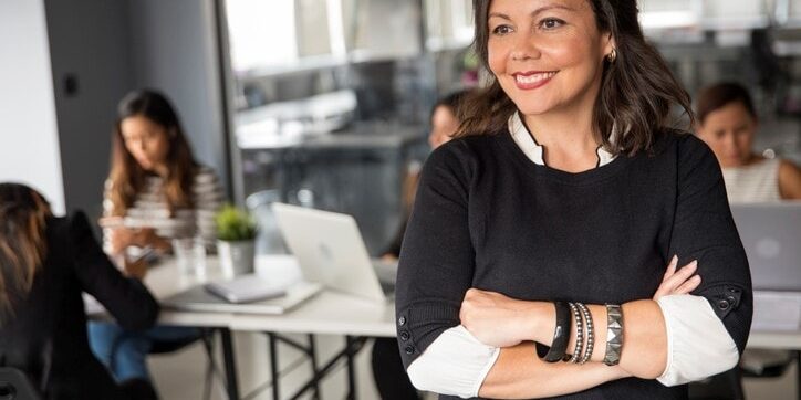 Smiling woman in a black sweater stands with arms crossed in front of a desk where four people work on laptops in a modern office.