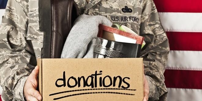 Person in military uniform holding a donation box with clothes and household items, in front of a U.S. flag.