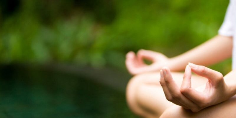 Person sitting cross-legged, meditating with hands resting on knees in a lush green outdoor setting.
