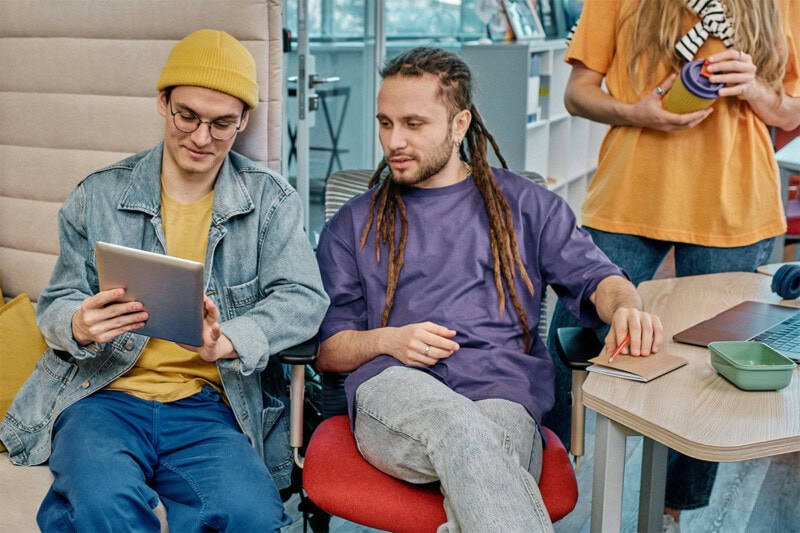 construct-check Two people are seated and looking at a tablet. One wears a yellow beanie and denim, the other a purple shirt. A third person stands nearby with a notebook and drink. Laptop on the table.