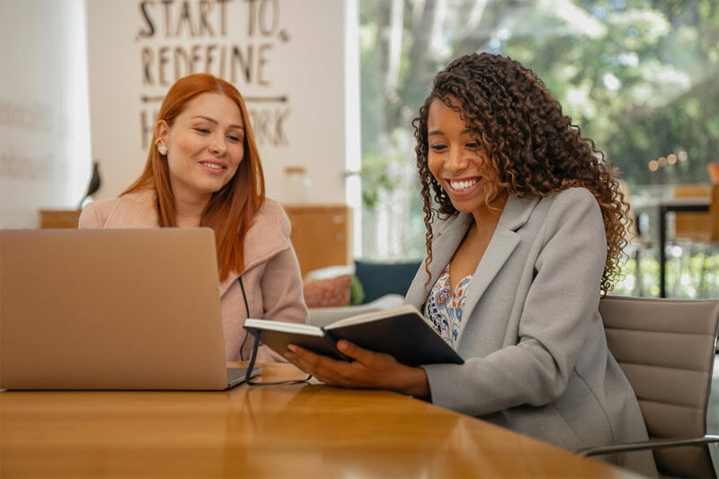 construct-spend-policy Two women sitting at a table with a laptop, one holding an open notebook and smiling, in a brightly lit room with a sign in the background.
