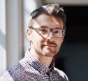 A man with short hair, glasses, and a beard wearing a checkered shirt stands indoors with sunlight coming through a window.