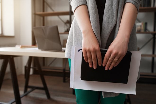 Person standing in an office, holding a book and papers. A laptop is open on the desk in the background.