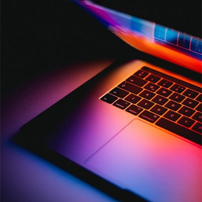 Partially open laptop with a colorful screen reflecting vibrant hues of blue, pink, and orange on the keyboard and surrounding surface.