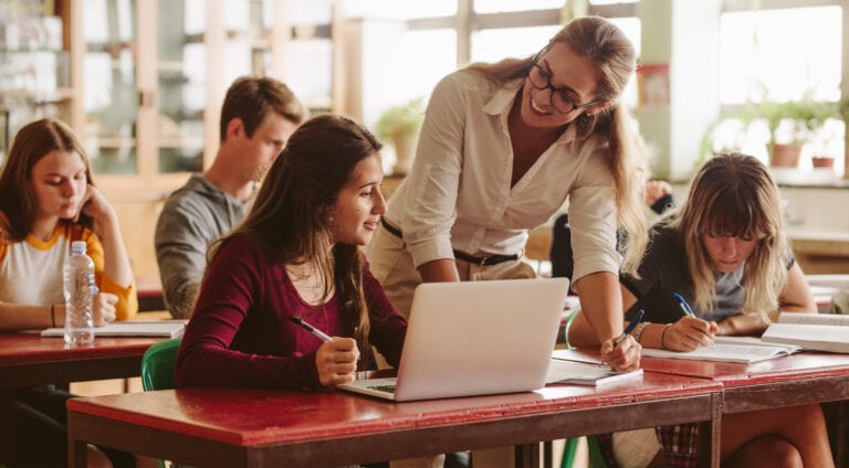 A teacher assists a student with a laptop in a classroom, while other students work at desks.