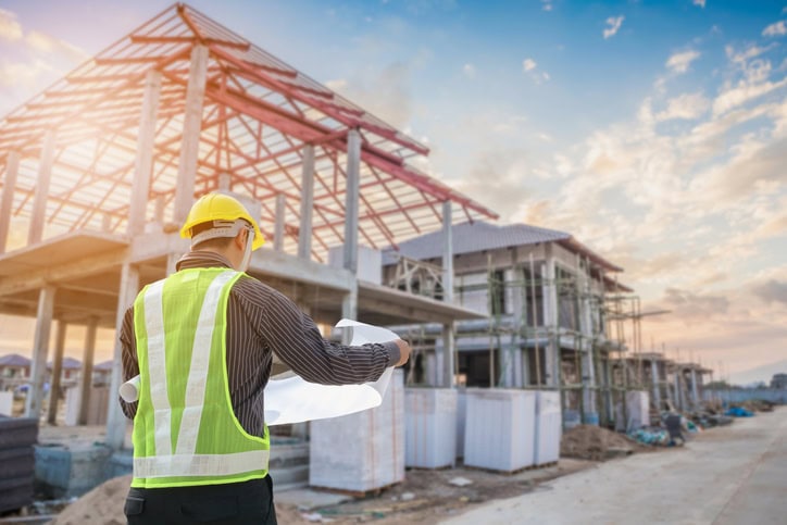 A construction worker in a yellow hard hat and safety vest reviews blueprints at a building site with partially completed structures and scaffolding under a blue sky.