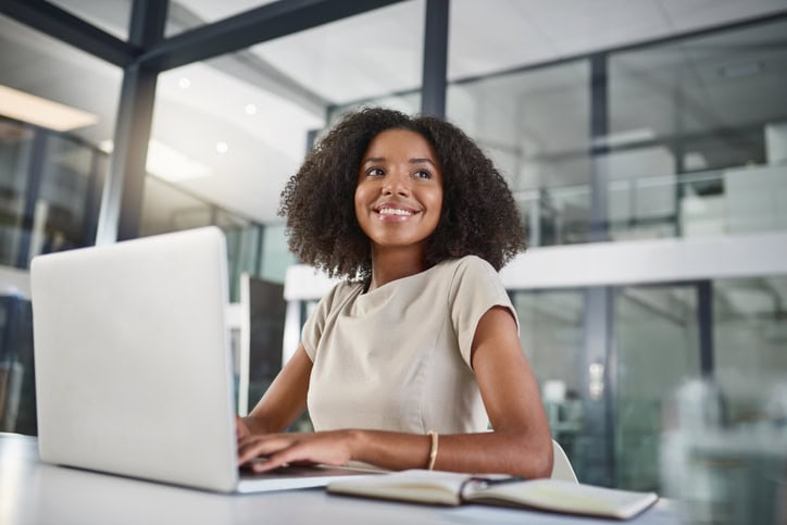 A woman sits at a desk in an office, smiling as she reviews travel expense management details on her laptop with an open notebook beside her.