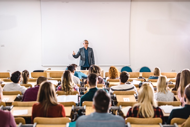 A man stands at the front of a lecture hall, addressing a group of seated students.