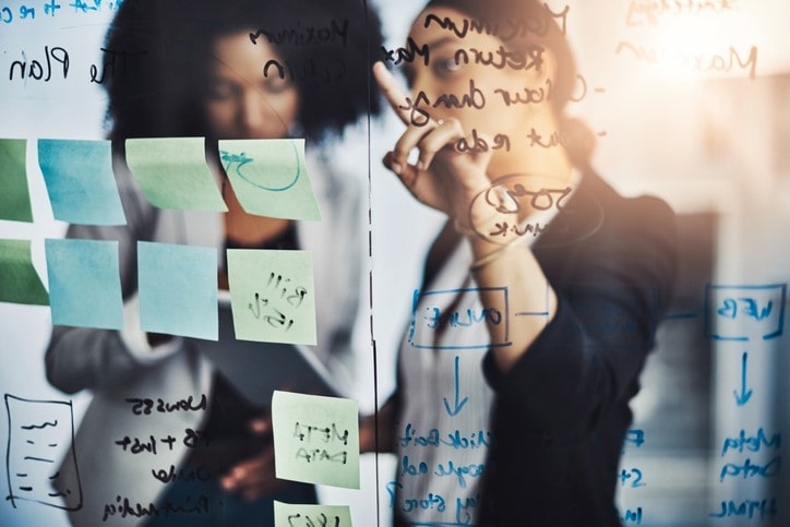 iStock-1132763550.jpg Two people brainstorming, writing on a transparent board with sticky notes and diagrams, in a well-lit room.
