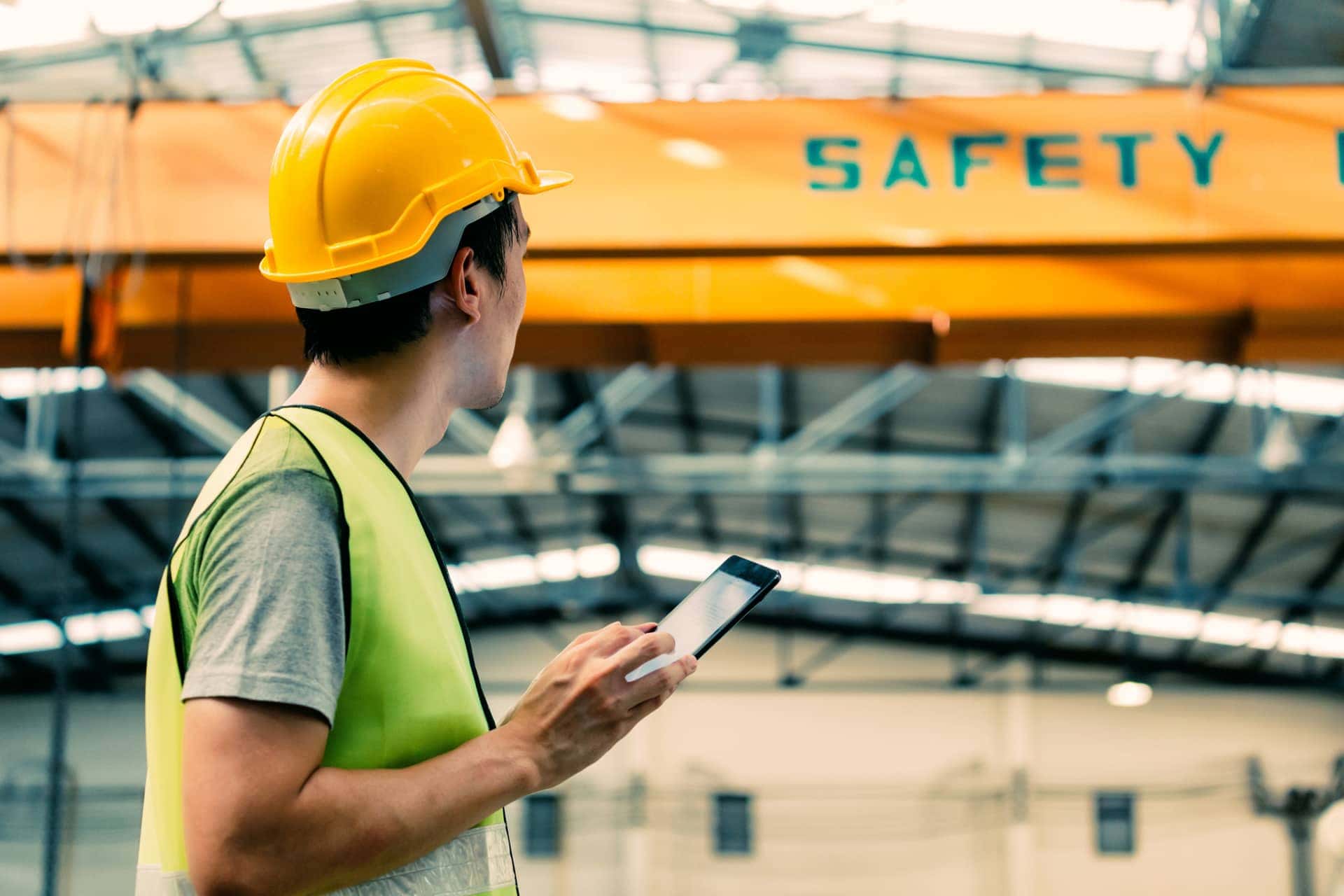 Worker in a yellow hard hat and safety vest holds a tablet, looking at overhead crane in an industrial setting.