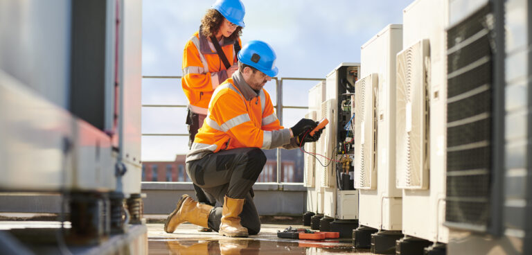 Two workers in safety gear inspect rooftop HVAC units. The man kneels with tools, the woman stands with a clipboard.