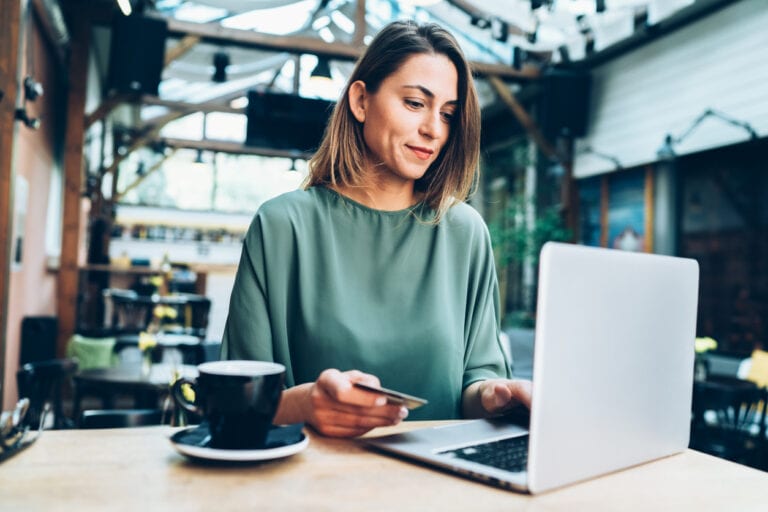 A woman in a green shirt is sitting at a table in a café, holding a card and using a laptop. A cup is on the table beside her.