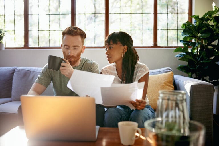 A man and woman sit on a couch, reviewing documents and drinking from mugs, with a laptop on the table in front of them.