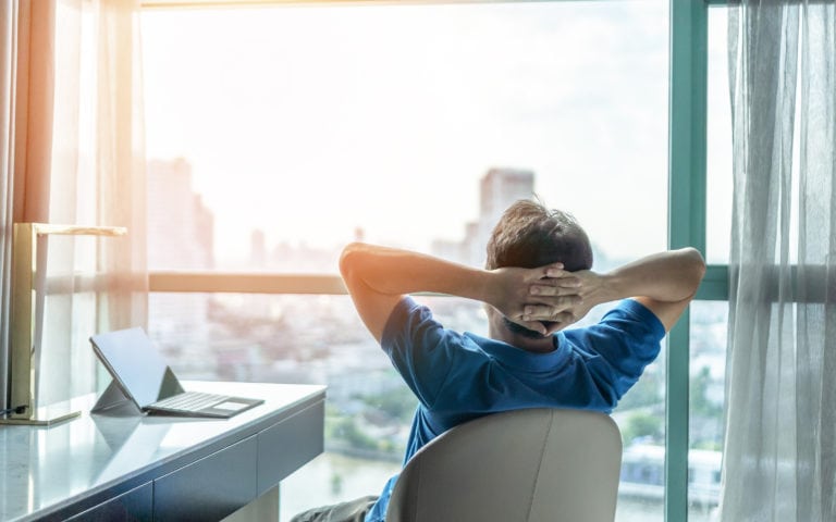 Person relaxing in a chair, facing a large window with a city view. A laptop is on the desk nearby.