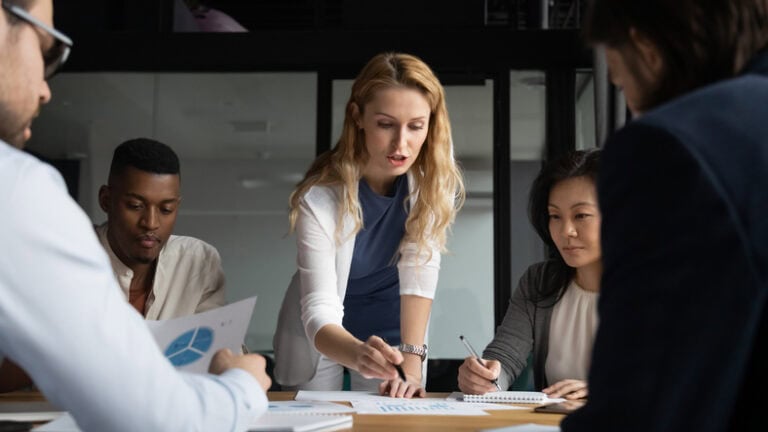 Young businesswoman explaining research results in graphs to colleagues. A group of five people are in a meeting room discussing papers with charts on a table. A woman in the center is pointing at a document.