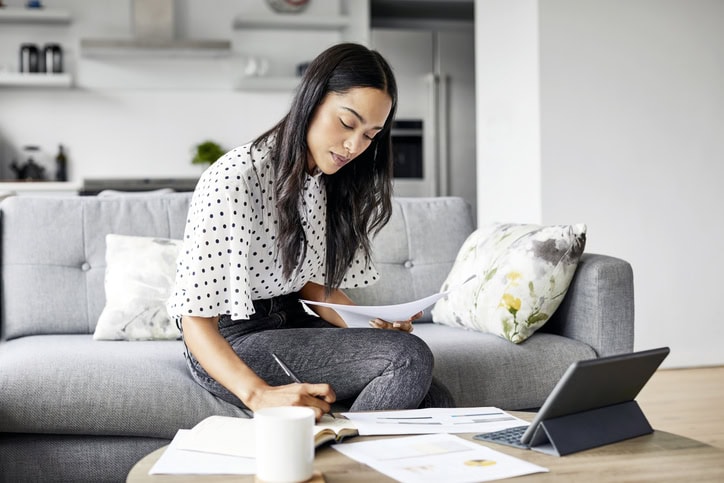 A woman sits on a sofa, working with papers and a tablet on a coffee table in a modern living room.