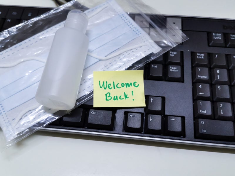 Welcome note with hand sanitizer and mask on work keyboard; Back to work note with alcohol gel to prevent coronavirus / infection