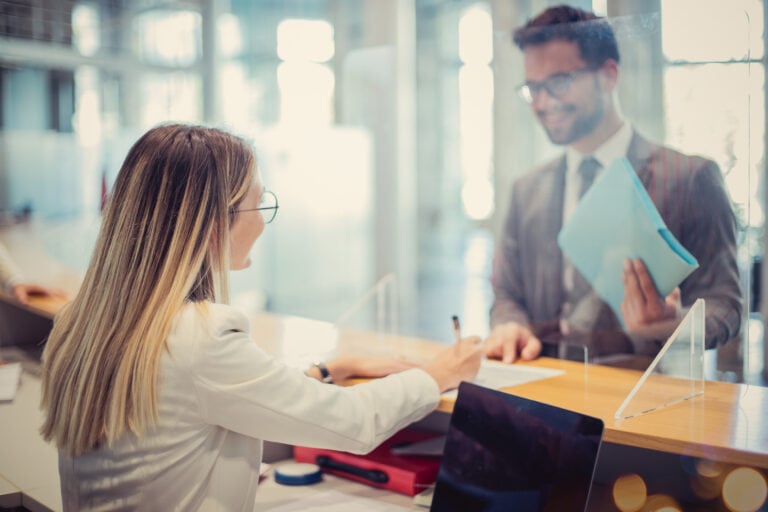 A woman signs a document at a reception desk while a man in a suit stands on the other side holding a blue folder.