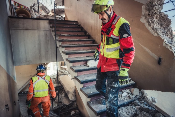 Two rescue workers in helmets and protective gear carefully navigate a debris-covered staircase in a building ravaged by the hurricane.