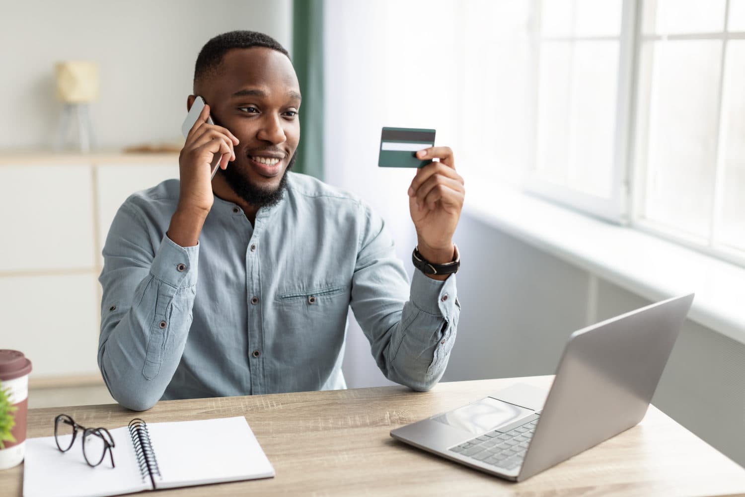 African Businessman Holding Credit Card Talking On Phone In Office Man in a light blue shirt sits at a desk, holding a credit card and talking on a phone, with a laptop and notepad in front of him.