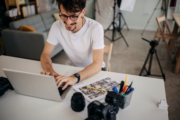 A man in a white t-shirt works on a laptop at a desk with cameras, lenses, and pens. A photo sheet and tripod are nearby.