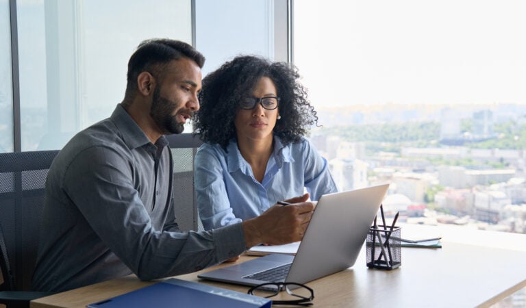 Two people sit at a table in an office, looking at a laptop screen, with a view of a cityscape visible through large windows.