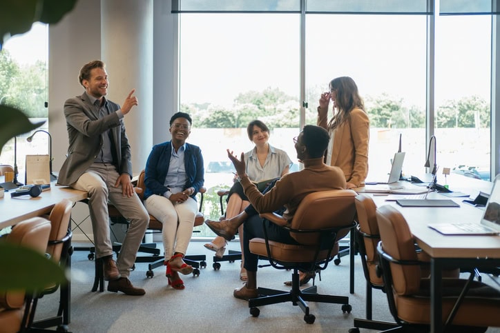 A diverse group of five people are having a lively discussion in a bright office space, seated around a table with laptops and papers.
