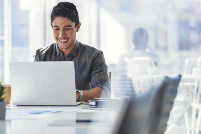 A person sits in a bright office, smiling and working on a laptop.