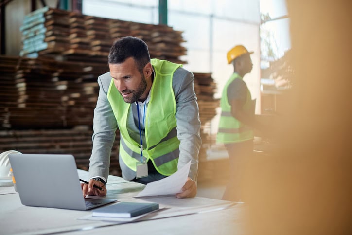 A man in a green safety vest works on a laptop while holding papers in a warehouse. Another person in a hard hat is visible in the background.