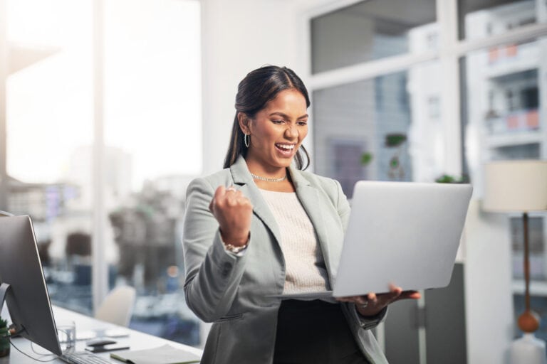 A person in business attire smiles and pumps their fist while looking at a laptop in a bright office.