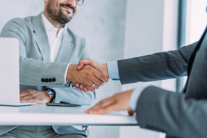 Successful partnership Two people in business attire shaking hands across a table, with a laptop and a smartphone nearby.