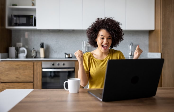 A person with curly hair looks excited while sitting at a kitchen table with a laptop and a coffee mug.