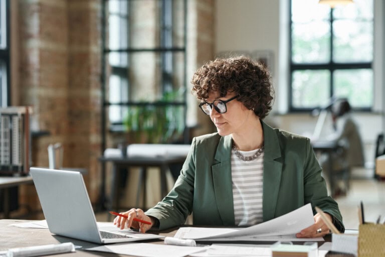 A person with curly hair and glasses works at a desk, analyzing financial statements on a laptop screen amidst an accounting talent shortage, in a well-lit office with large windows.