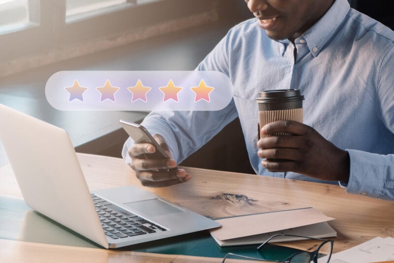 Closeup cropped portrait of satisfied businessman consumer with phone giving five stars feedback on food service holding plastic coffee cup in hands sitting at office workplace in front of laptop