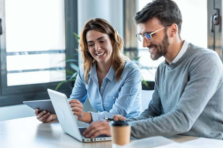 Smart business people working with plans while talking of their new design business in the office Two people working at a desk with a laptop and tablet, smiling and appearing collaborative. A coffee cup is on the desk.