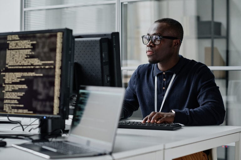 A person wearing glasses works at a desk with multiple computer monitors displaying code.