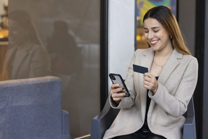 A woman in a beige blazer sits and smiles at her phone while holding a prepaid card.
