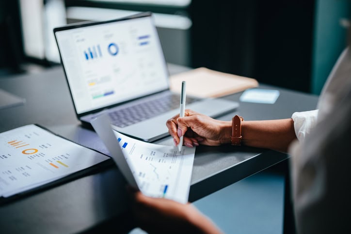 Close Up Photo Of Woman Hands Writing Report On A Paper  In The Cafe Person analyzing graphs on a tablet and laptop at a desk.