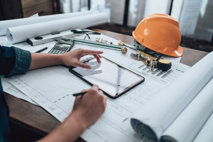 Person working on architectural plans with a tablet, surrounded by drafts, calculator, helmet, and drawing tools on a desk.