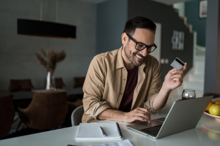 A man sitting at a kitchen counter holds a corporate card and smiles while using a laptop.