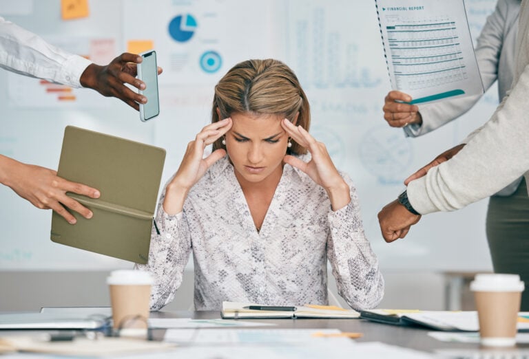 Woman, headache and office with stress, multitasking deadline or tired of project management target. Corporate executive, documents or email on schedule, anxiety and burnout of mental health by desk A stressed woman sits at a desk with hands on temples, surrounded by people presenting documents, a tablet, and a phone. Coffee cups and paperwork are on the table.