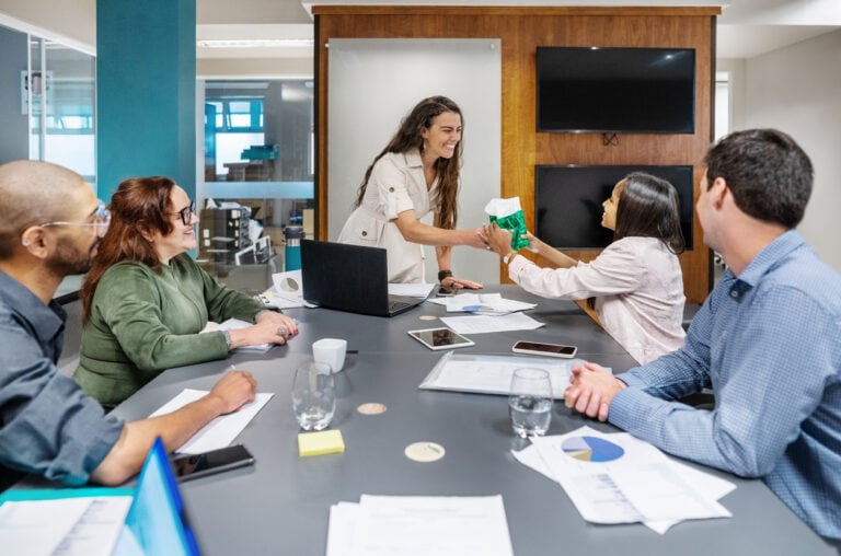 Five people in a meeting room, seated around a table with documents and laptops, while one person hands a gift bag to another.