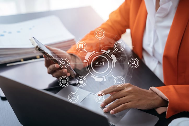 Modern technology and networking concept, woman using laptop, tablet and smartphone with virtual screen API icon Software development tool. API Application Programming Interface. Person in an orange blazer uses a smartphone and laptop. A virtual diagram labeled "API" features icons like a lock, gear, and cloud, symbolizing technology and connectivity concepts.