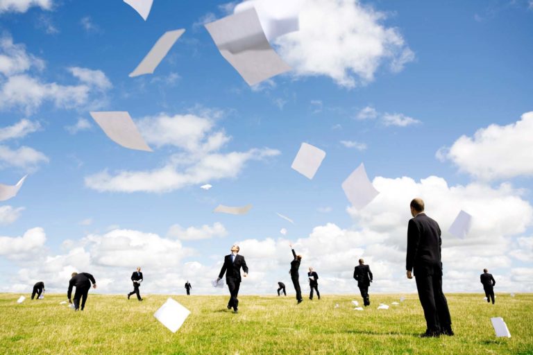 Men in suits stand on a grassy field as sheets of paper float through the air under a partly cloudy sky.