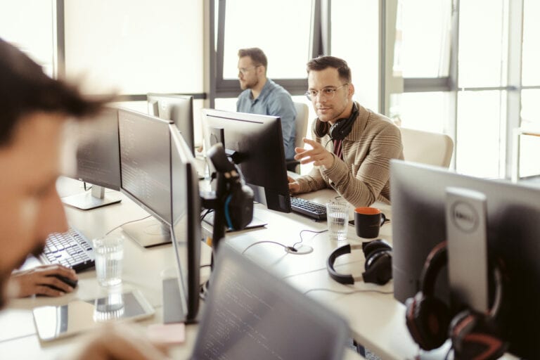 Office scene with several people working at computers. One person is gesturing while speaking, and others are focused on screens. Headphones, keyboards, and coffee mugs are visible.