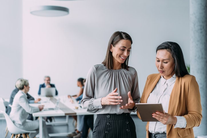 Business people in the office. Two women in a modern office discuss something while one holds a tablet. Background shows colleagues working at a table.